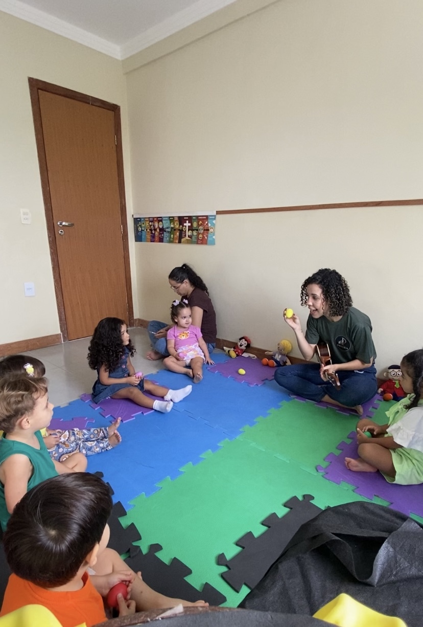 Turma de musicalização infantil sentada no tapete acompanhando a professora com bolinhas coloridas