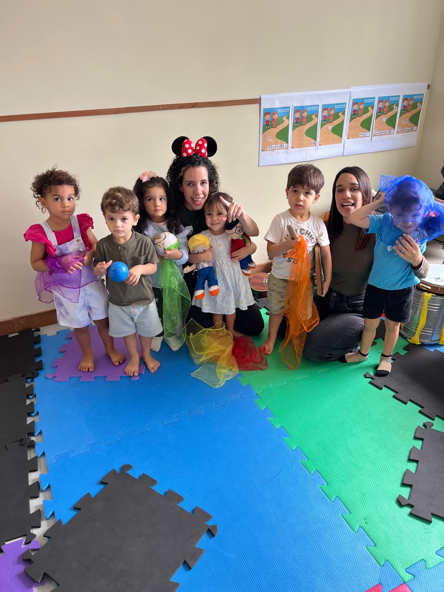 Crianças com lenços coloridos, brinquedos e instrumentos ao lado das professoras após a aula de musicalização