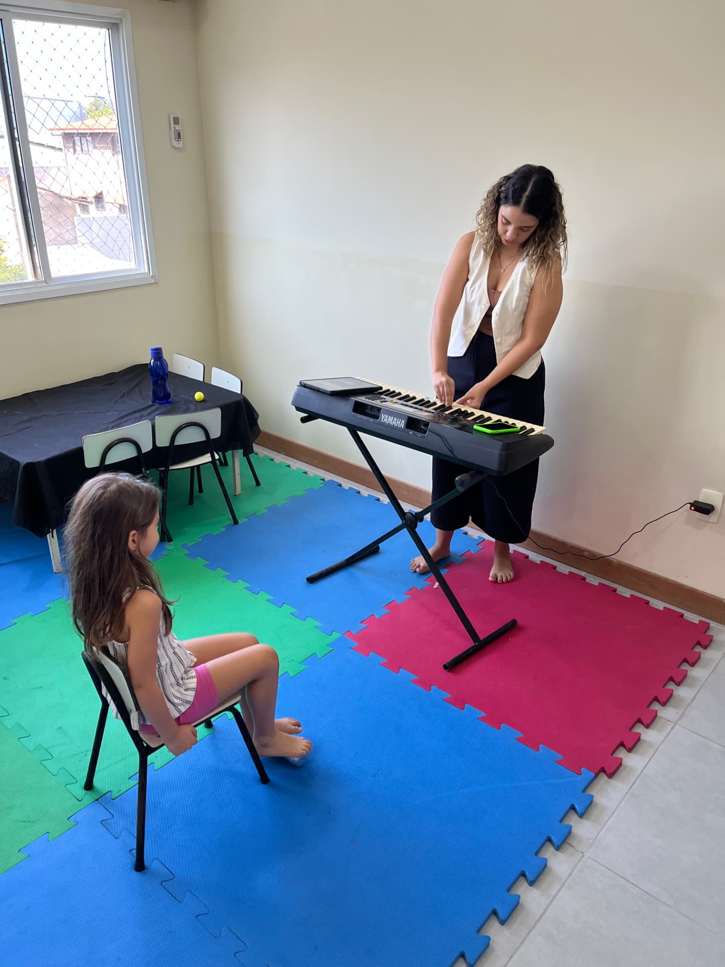 Professora ao teclado conduzindo aula individual de canto para uma criança