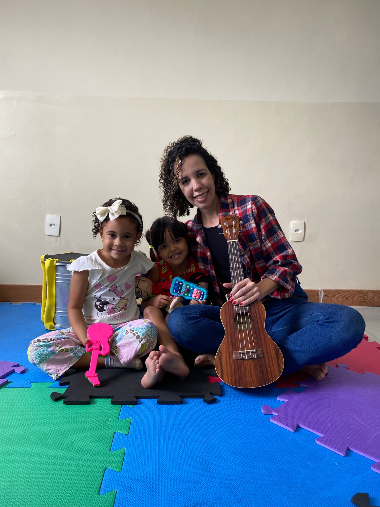 Professora do Projeto PHIARA sentada no tapete com duas crianças, segurando um ukulelê durante a aula