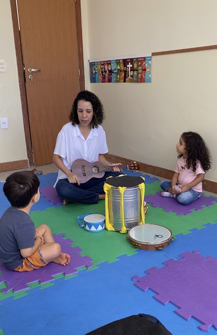Professora tocando ukulelê em roda com duas crianças e instrumentos de percussão no chão